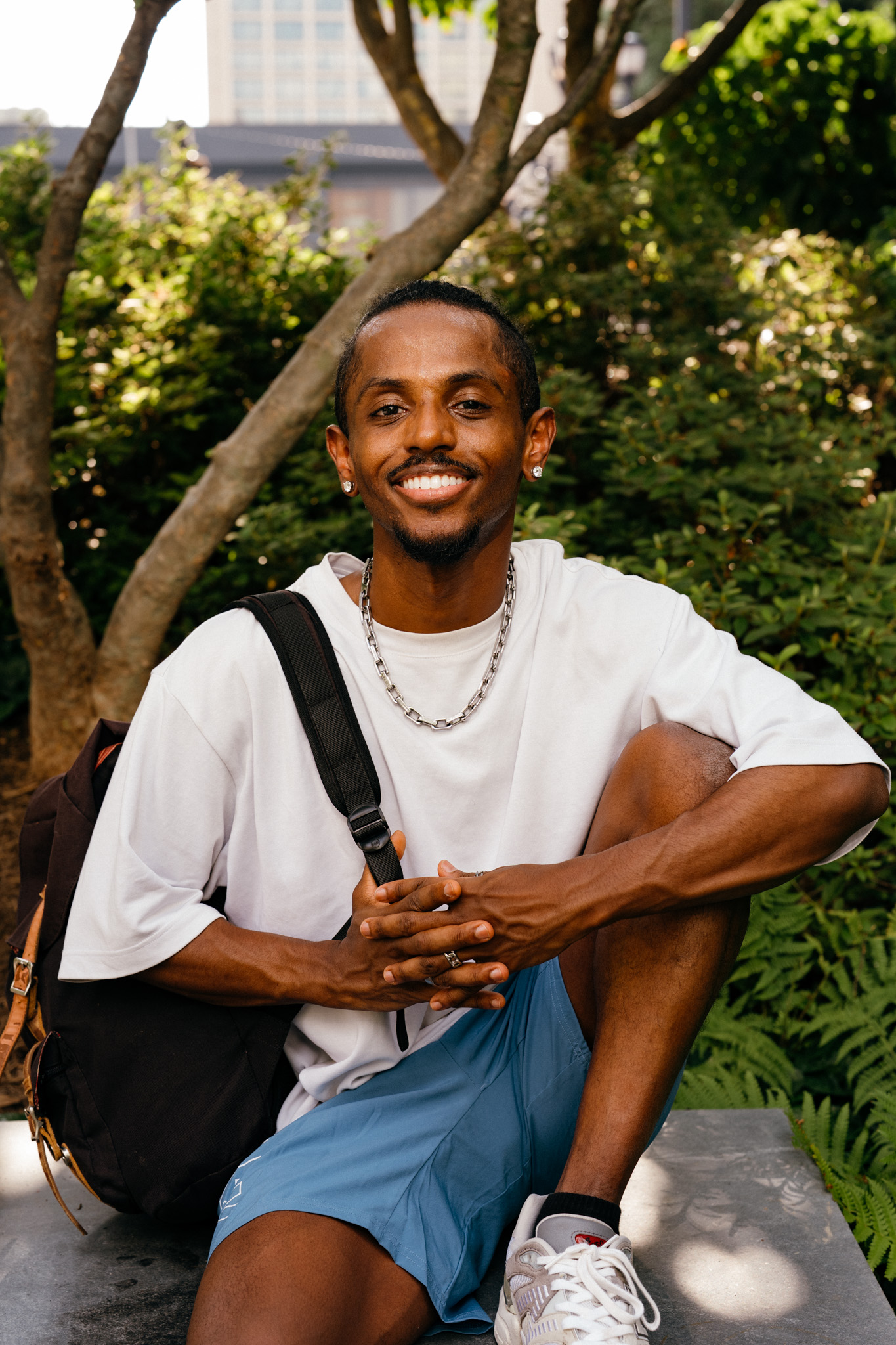 Meni seated with backpack in warm natural light with trees behind him, Crystal City, July 2026.