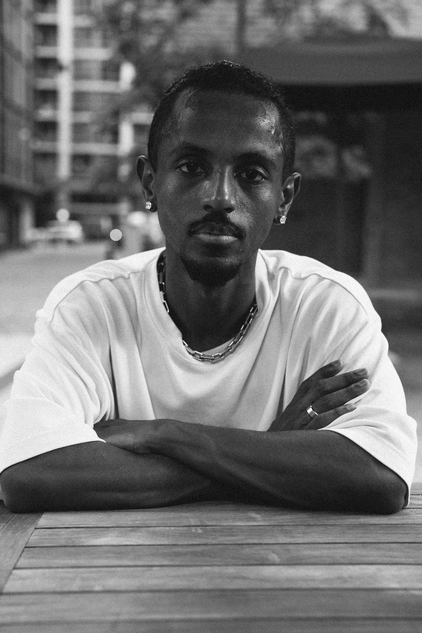 Meni seated at an outdoor cafe table, arms crossed, looking directly at camera, black and white portrait, Crystal City, July 2026.