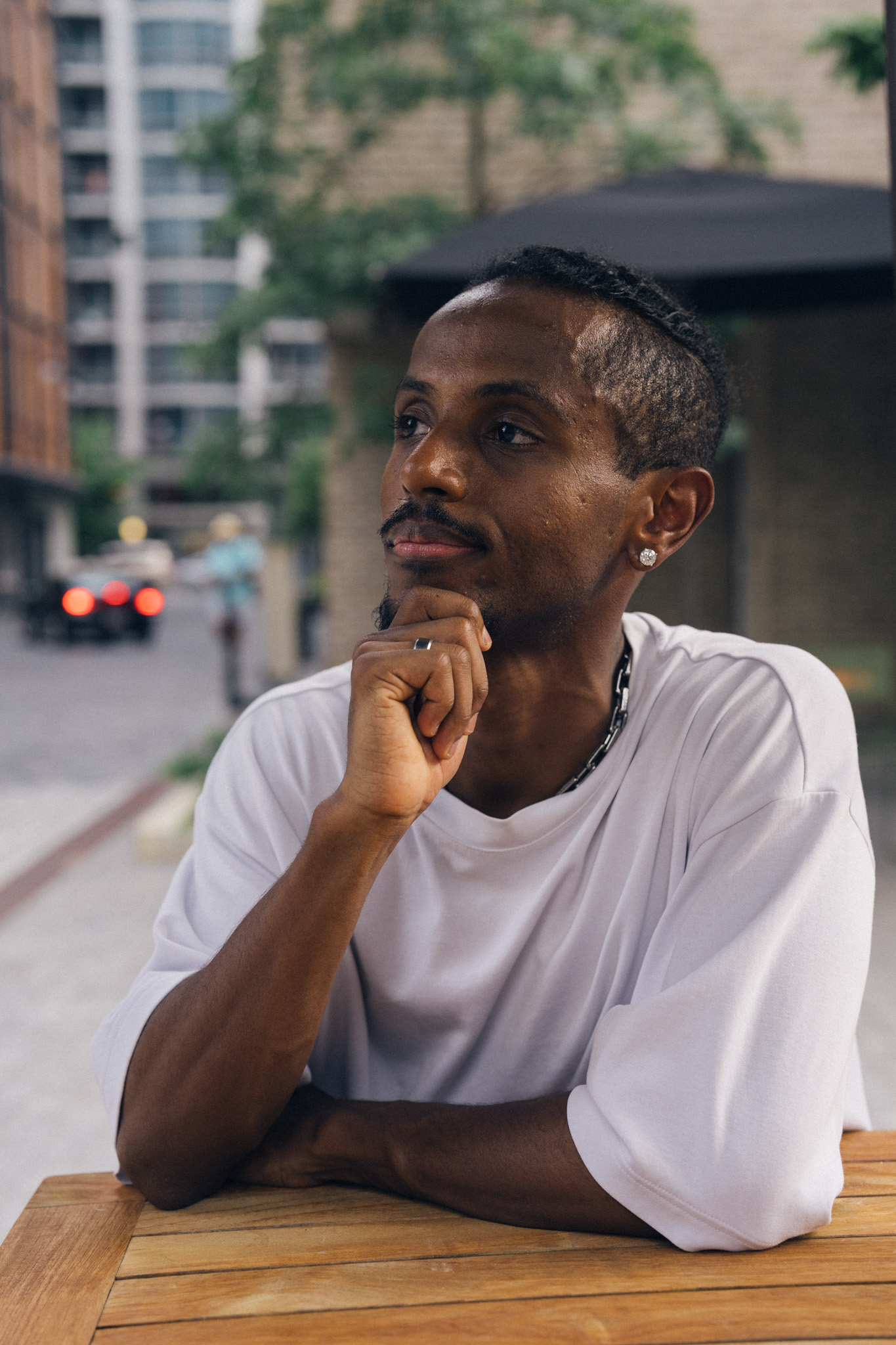 Meni seated at an outdoor cafe table, hand on chin, looking away contemplatively, Crystal City, July 2026.