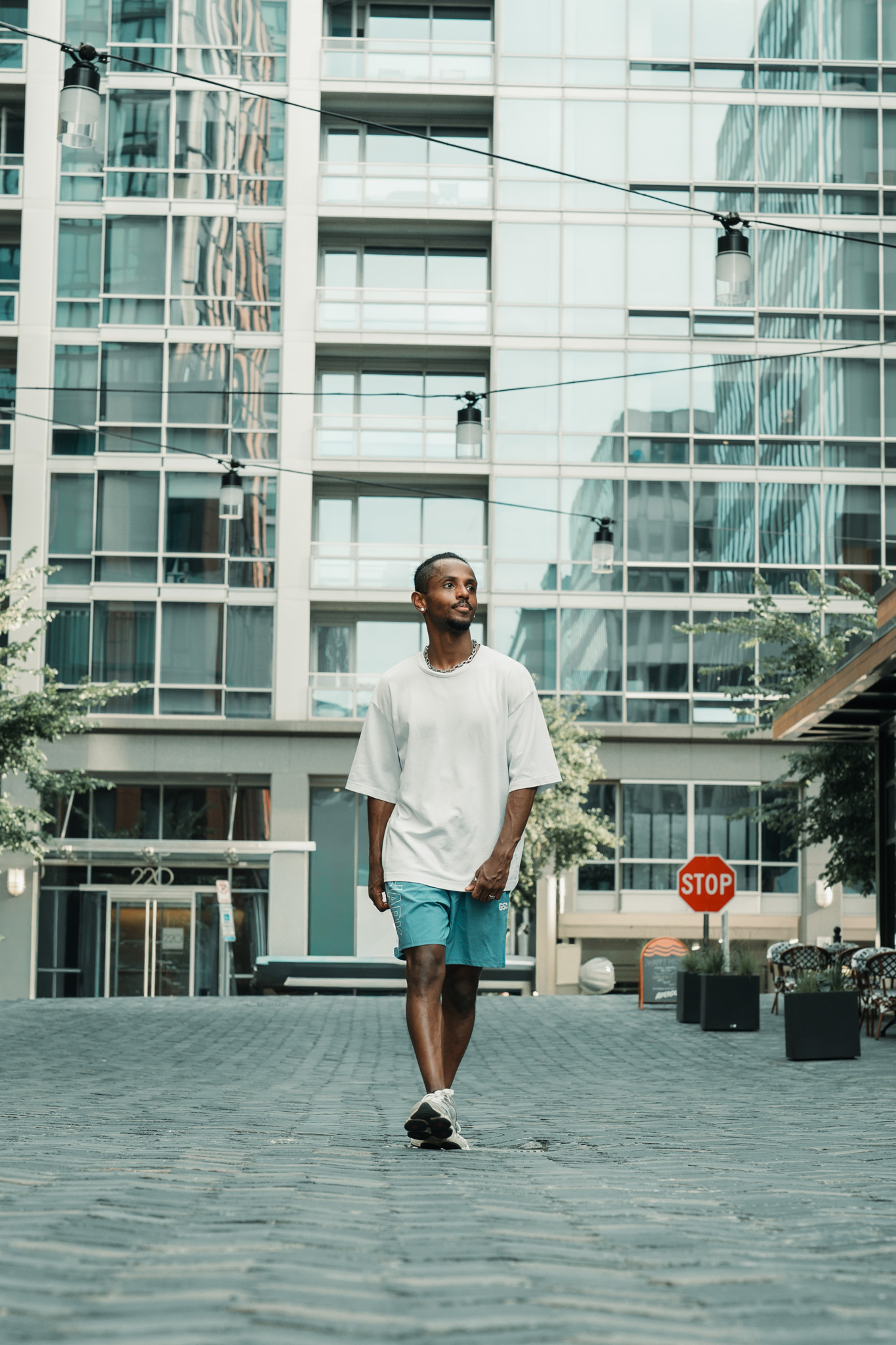 Meni full body walking with a glass high-rise building behind him, Crystal City courtyard, July 2026.